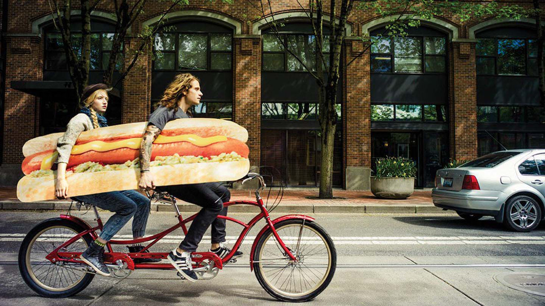 大きなホットドックの看板を二人で脇に抱えながらタンデム自転車を漕いでいる様子