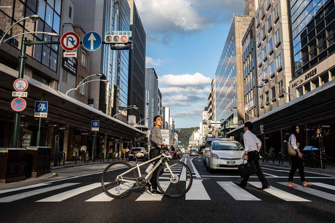 青空が反射しているビルの窓と空が印象的な道路の横断歩道をクロスバイクを押して歩く写真