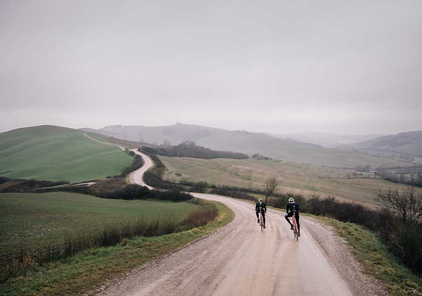 広大な緑の丘にクネクネと続く砂利道を走るグラベルロードの二人の写真