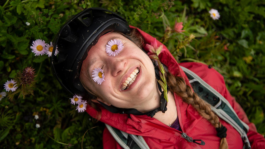 草むらに寝転び、淡いピンクの花を目の上にのせて笑う女性の写真