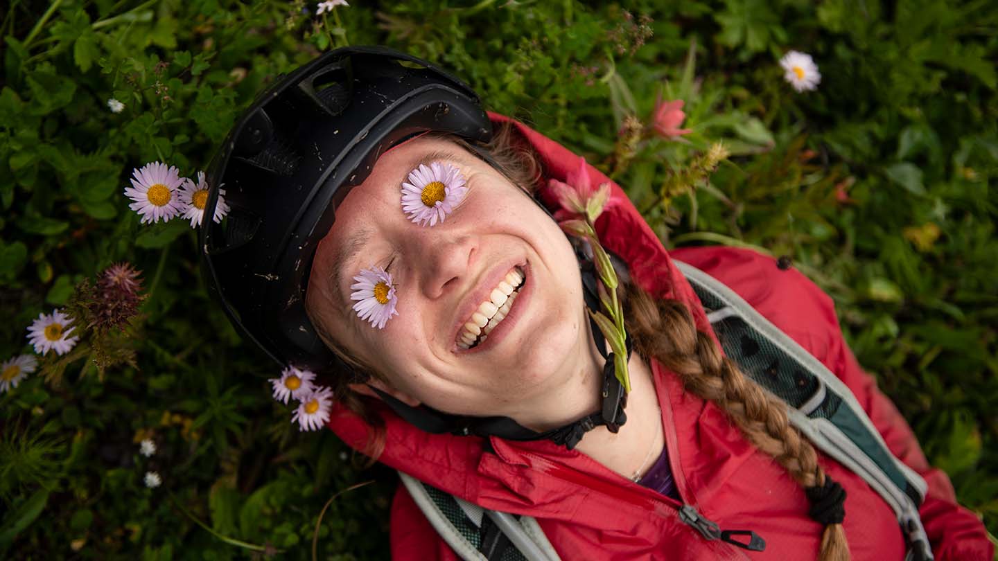 野の花が咲く地面に寝そべり両目の上に花をのせて笑うサイクリストの写真