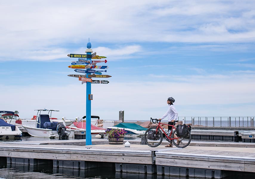 旅先のヨットハーバーで多数の行き先を示す看板を見るパニアバッグをつけたロードバイクサイクリストの写真