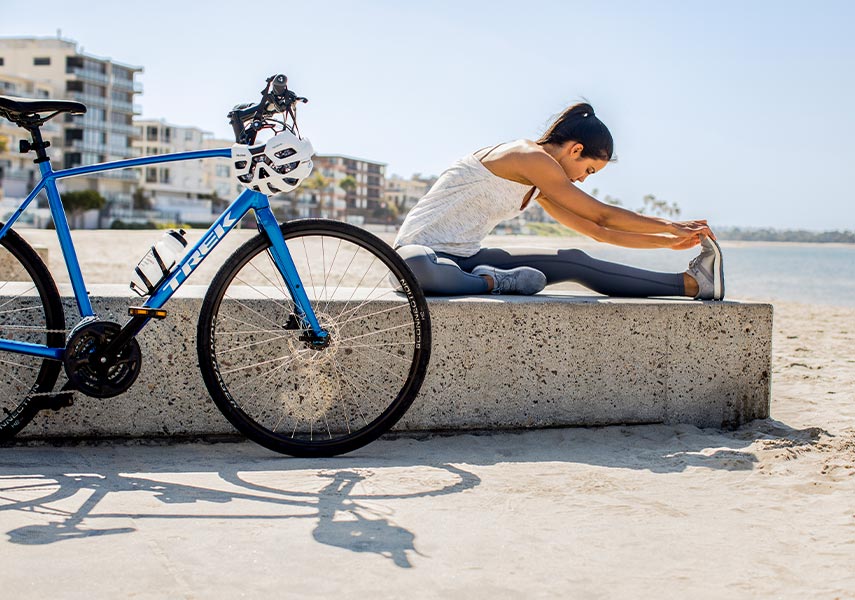 白い砂浜で青いクロスバイクの脇でストレッチをする女性サイクリストの写真